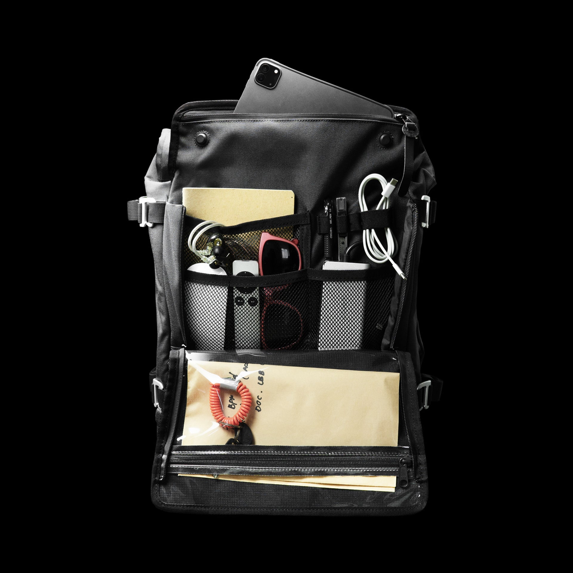 The Peloton Asphalt S opened flat on black, organiser panel loaded with a tablet, cables, sunglasses, notebook, keys and documents in mesh and clear-window pockets.