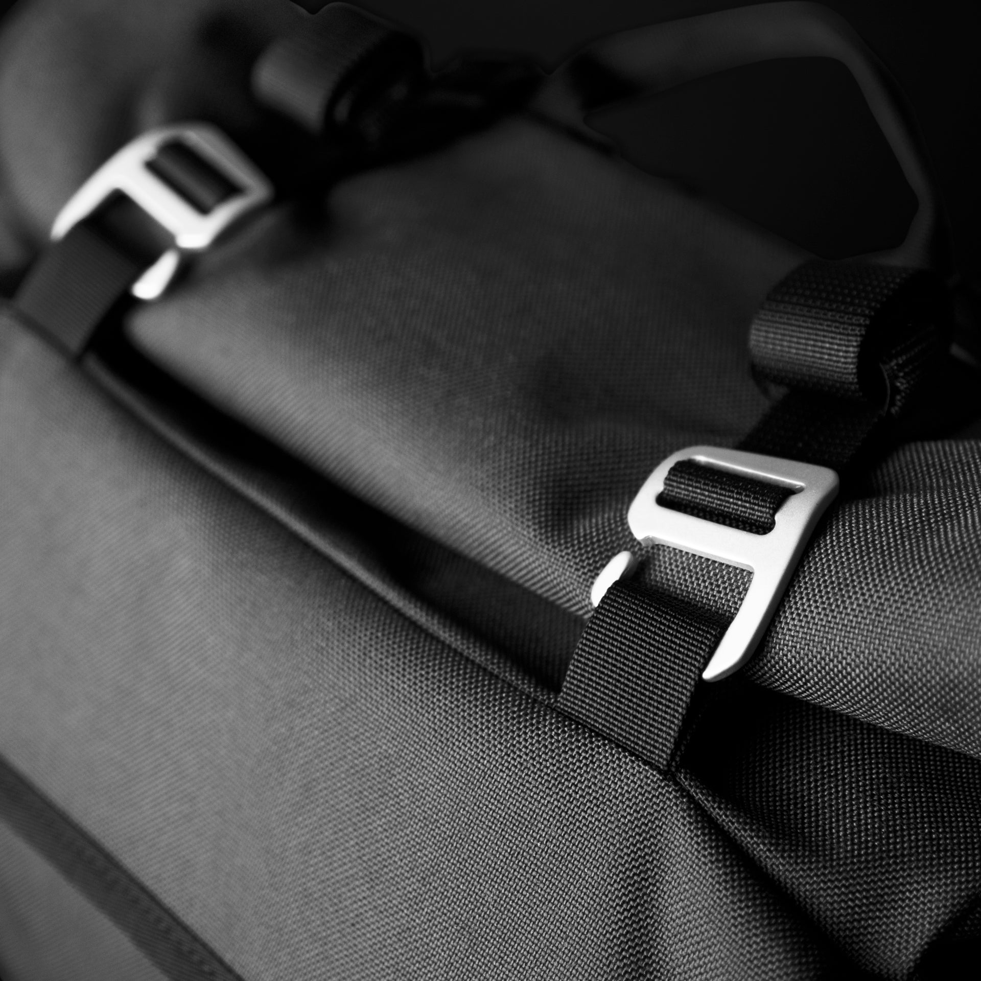 Macro detail of the Asphalt S machined aluminium G-hook buckle threaded through a black nylon compression strap against ballistic weave.