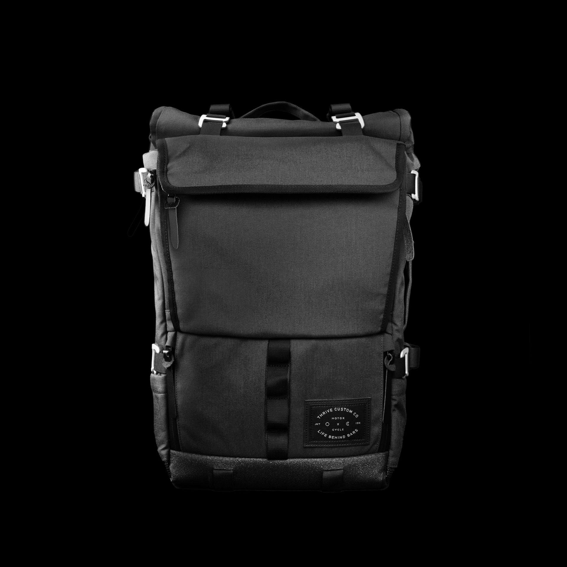 The Peloton Asphalt S roll-top backpack, front view on black, showing the flap closure, dual compression straps and a Thrive Custom Co × Life Behind Bars woven patch.